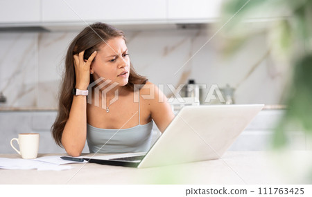 Young puzzled woman working on laptop in kitchen Young puzzled woman working on laptop in kitchen 111763425