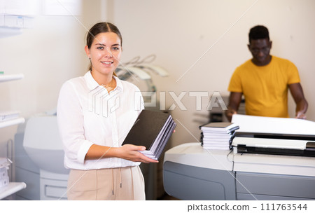 Cheerful young woman in white shirt smiling at the camera and holding new planners in the printing house 111763544
