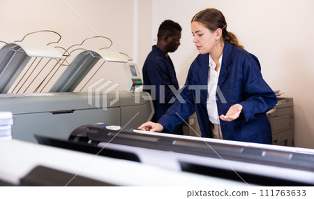 Focused young woman in uniform setting up the plotter before starting work in the printing house 111763633