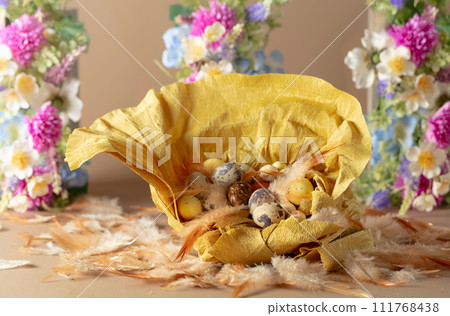 Crepe paper nest with easter eggs, feathers, and spring flowers on a beige background. 111768438