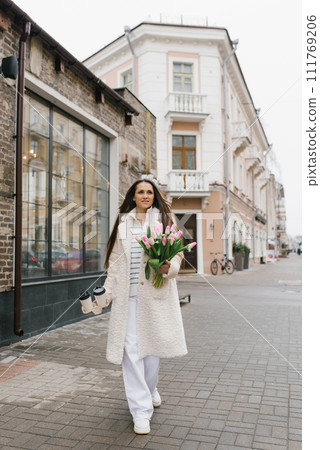 Young stylish woman in a light-colored coat walks down a city street with a paper cup of coffee or tea and a bouquet of spring tulips Young stylish woman in a light-colored coat walks down a city street with a paper cup of coffee or tea and a bouquet of spring tulips 111769206