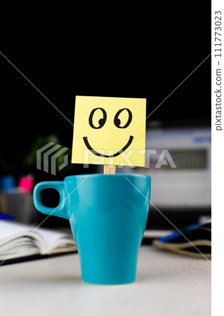 Smiling face drawn on sticky note inside a cup of coffee at the office desk. Every morning wake up with the smile Smiling face drawn on sticky note inside a cup of coffee at the office desk. Every morning wake up with the smile 111773023