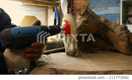 A grinding machine with iron bristles in the hands of a craftsman polishing a piece of whole wood material in the workshop, the woodworker cleans a metal brush on a drill of uneven natural wood A grinding machine with iron bristles in the hands of a craftsman polishing a piece of whole wood material in the workshop, the woodworker cleans a metal brush on a drill of uneven natural wood 111773092