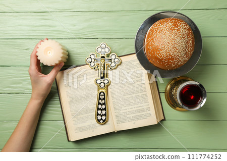 Candle in hand, bread, cup with wine and cross on book on green wooden background, top view Candle in hand, bread, cup with wine and cross on book on green wooden background, top view 111774252