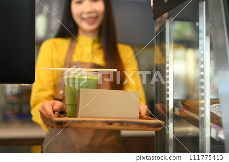 Smiling waiter in apron serving cold drink with paper straw and cake in a cardboard takeaway box 111775413