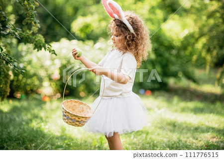 Easter egg hunt. Girl child Wearing Bunny Ears Running To Pick Up Egg In Garden. Easter tradition. Baby with basket full of colorful eggs. Easter egg hunt. Girl child Wearing Bunny Ears Running To Pick Up Egg In Garden. Easter tradition. Baby with basket full of colorful eggs. 111776515
