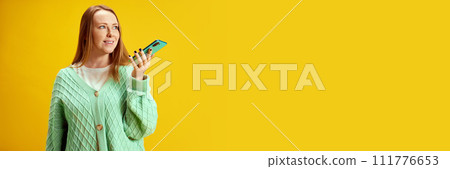 Banner. Young woman using smartphone and speaking by cellular connection against yellow studio background with negative space against studio background. 111776653