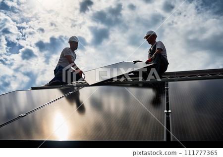 Workers building solar panel system on rooftop of house. Two men installers in helmets installing photovoltaic solar module outdoors. Alternative, green and renewable energy generation concept. Workers building solar panel system on rooftop of house. Two men installers in helmets installing photovoltaic solar module outdoors. Alternative, green and renewable energy generation concept. 111777365
