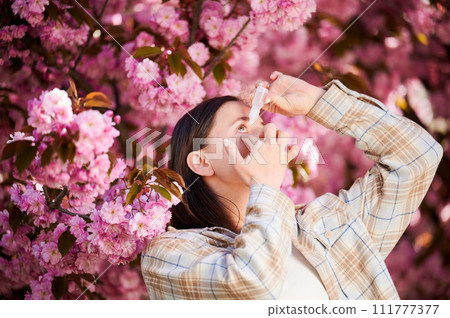 Woman allergic using medical eyes drops, suffering from seasonal allergy at spring in blossoming garden. Young woman treating eyes in front of blooming tree outdoors. Spring allergy concept. 111777377