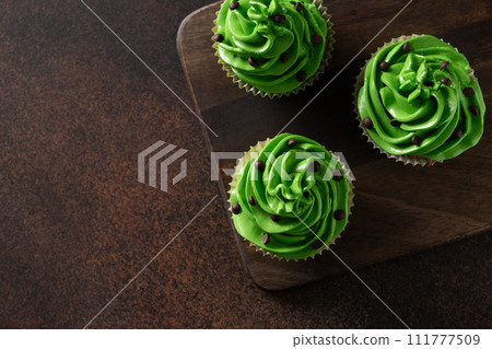 St. Patrick's Day chocolate cupcakes with green whipped cream decorated shamrock and clover and chocolate sprinkles on brown background for Close up. St. Patrick's Day chocolate cupcakes with green whipped cream decorated shamrock and clover and chocolate sprinkles on brown background for Close up. 111777509