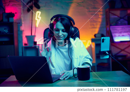 Online education. Young woman sitting in room with neon lights, with laptop, leading online video lesson, stream. 111781197