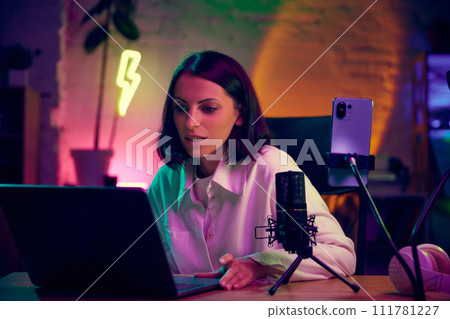 Young woman, blogger sitting in modern room with neon lights, working with laptop, leading online video lesson. Online education 111781227
