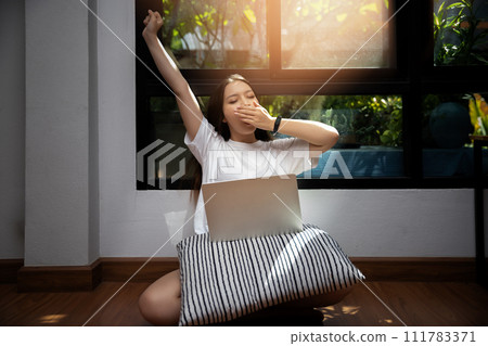 Young asian woman open mouth yawning while sitting on the floor in living room. Studying online at home. Distance learning. Remote work place. Quarantine, coronovirus covid-19. Young asian woman open mouth yawning while sitting on the floor in living room. Studying online at home. Distance learning. Remote work place. Quarantine, coronovirus covid-19. 111783371