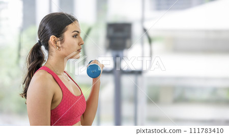 Young Focused female athlete performing a bicep curl with a blue dumbbell in a bright gym. 111783410