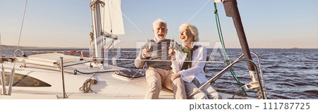 Happy senior couple sitting on the side of sail boat or yacht deck floating in sea. Man and woman drinking wine or champagne and laughing Happy senior couple sitting on the side of sail boat or yacht deck floating in sea. Man and woman drinking wine or champagne and laughing 111787725