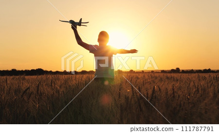 Cheerful boy teen running with plane toy on autumn wheat field at cinematic sunset sunrise back view closeup. Active male kid playing flying airplane plaything enjoy freedom happy childhood at meadow 111787791