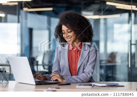 Smiling black woman in eyeglasses using pc while working by equipped table during daytime. Successful data analyser doing online research for making development strategy and enjoying modern device. 111788045