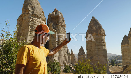 A man in a yellow T-shirt and baseball cap takes a picture of the mountains with his phone. Love Valley in Cappadocia. A blogger shoots a story on a smartphone about cliffs in Turkey. A man in a yellow T-shirt and baseball cap takes a picture of the mountains with his phone. Love Valley in Cappadocia. A blogger shoots a story on a smartphone about cliffs in Turkey. 111788404
