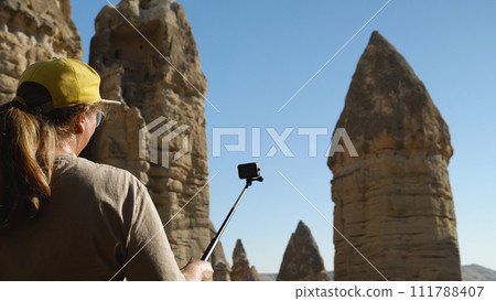 Woman filming herself on action camera against a backdrop of beautiful sandstone cliffs. A girl records a video for her blog from her holiday in Turkey. Love Valley in Cappadocia. 111788407