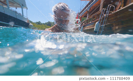 A man emerges from the sea and shakes off the water with a movement of his head. Two pleasure boats or yachts are anchored next to him. Underwater photography, Concept of a summer trip. 111788409