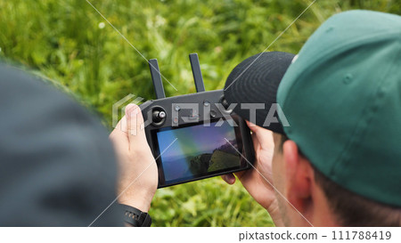 Close-up of drone pilot's hands controlling the quadrocopter remote control while moving. View of hands and screen. The screen shows the image from the camera of the UAV drone. Close-up of drone pilot's hands controlling the quadrocopter remote control while moving. View of hands and screen. The screen shows the image from the camera of the UAV drone. 111788419