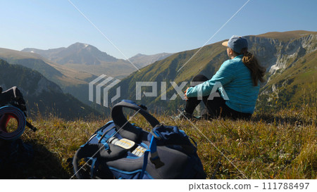 A female traveller sits on the edge of a cliff and looks at a beautiful mountain landscape. In the foreground is a hiking rucksack. Summer hiking in the mountains. 111788497