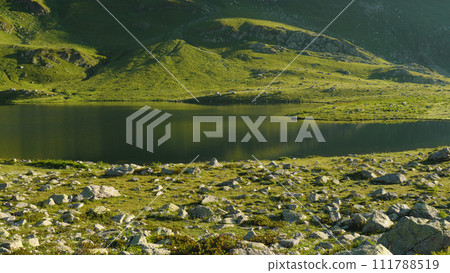 A beautiful green mountain lake. Rocks are scattered in the foreground. Green grass grows around the lake. A picturesque mountain lake in the morning. The beauty of the wildlife. 111788519