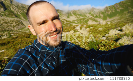 The man's face is red from tanning. A bearded Caucasian man with a short haircut is smiling as he looks into the camera. Against a background of beautiful mountains. A travelling blogger. 111788520