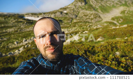 The face of a bearded middle-aged Caucasian man with a short haircut. Against the background of beautiful mountains and blue sky. A travelling blogger taking pictures of himself. 111788521