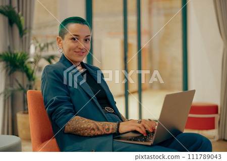 Smiling tattooed female freelancer with green short hair working on laptop while sitting in office Smiling tattooed female freelancer with green short hair working on laptop while sitting in office 111789043