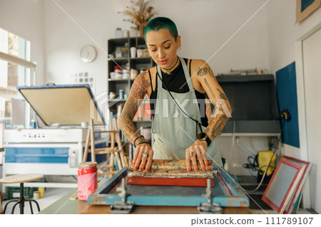 Female worker with squeegee is printing images on textile by silkscreen method in a design studio Female worker with squeegee is printing images on textile by silkscreen method in a design studio 111789107