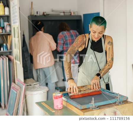 Female worker with squeegee is printing images on textile by silkscreen method in a design studio Female worker with squeegee is printing images on textile by silkscreen method in a design studio 111789156