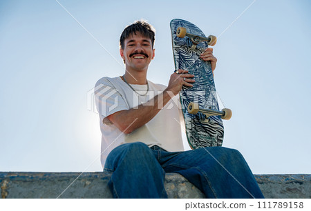 Low angle of smiling male skater in casual outfit sitting on ramp with skateboard against blue sky Low angle of smiling male skater in casual outfit sitting on ramp with skateboard against blue sky 111789158
