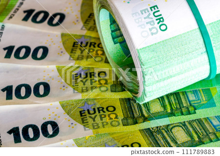 A roll of one hundred euro banknotes with a green rubber band on a background of 100 euro banknotes A roll of one hundred euro banknotes with a green rubber band on a background of 100 euro banknotes 111789883