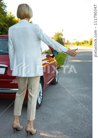 Woman standing near broken car on highway road asking for help from drivers raising her finger up Woman standing near broken car on highway road asking for help from drivers raising her finger up 111790347
