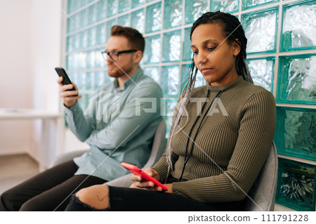 Two diverse young business people, hiring, using mobile phones in waiting room for networking, social media or communication at office. Group of employee workers wait in line with smartphone Two diverse young business people, hiring, using mobile phones in waiting room for networking, social media or communication at office. Group of employee workers wait in line with smartphone 111791228