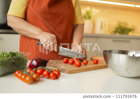 Hands of senior 60s female chopping fresh tomato on wooden board while preparing italian salad with vegetables  111791676