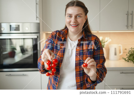 Smiling young woman holding tomato, standing at modern kitchen, chopping vegetables, preparing fresh healthy salad for dinner or lunch. Young Caucasian woman cooking at home, making breakfast 111791691