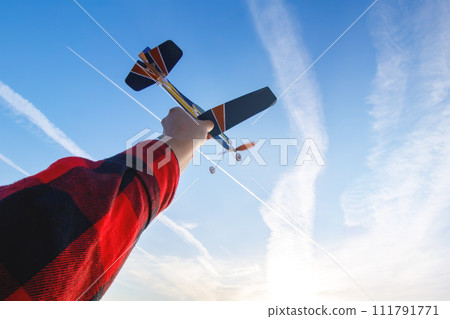 A model of a rubber-engined paper toy airplane in hand against a blue sky with clouds and a contrails (condensation trails, vapour trails) of a high-flying airliner. POV (First person view from eyes). A model of a rubber-engined paper toy airplane in hand against a blue sky with clouds and a contrails (condensation trails, vapour trails) of a high-flying airliner. POV (First person view from eyes). 111791771