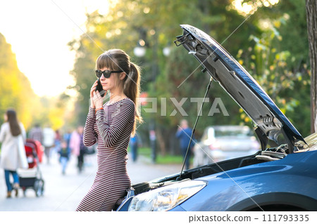 Upset young woman driver talking angrily on cell phone with assistance service near a broken car with open hood on a city street. Upset young woman driver talking angrily on cell phone with assistance service near a broken car with open hood on a city street. 111793335