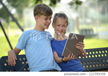 Two happy teenager children, girl and boy looking at screen of digital tablet, reading, studying or gaming together sitting on bench outdoors on summer sunny day 111793336