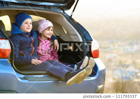 Two happy children boy and girl sitting together in a car trunk. Cheerful brother and sister hugging each other in family vehicle luggage compartment. Weekend travel and holidays concept. 111793337