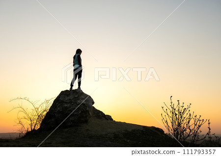 Silhouette of a woman hiker standing alone on big stone at sunset in mountains. Female tourist on high rock in evening nature. Tourism, traveling and healthy lifestyle concept. 111793357