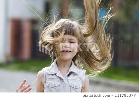 Pretty little preschool girl in sleeveless white dress with beautiful long blond hair blown by wind, funny toothless smile and spread arms on blurred background. Innocent happy childhood concept. Pretty little preschool girl in sleeveless white dress with beautiful long blond hair blown by wind, funny toothless smile and spread arms on blurred background. Innocent happy childhood concept. 111793377