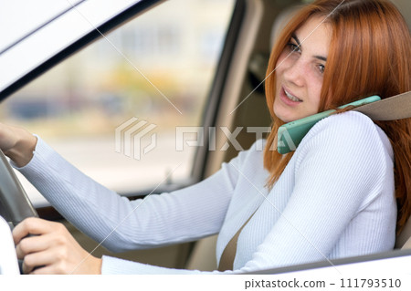 Happy redhead girl talking on her mobile phone behind the wheel driving a car. 111793510