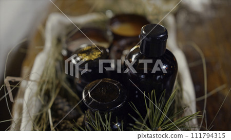 Aromatherapy table setting with perfumed candles, oil bottle and coniferous twig with cones creating mood. Video. Close up of decorative box with natural candles and oil on wooden table background. 111794315