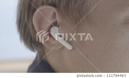 Close up of a man putting air pods in his ear. Stock. Side view of blond male face, he is putting white wireless earphone in his ear on blurred background. Close up of a man putting air pods in his ear. Stock. Side view of blond male face, he is putting white wireless earphone in his ear on blurred background. 111794463