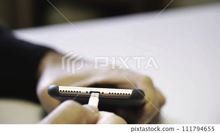 Close up of hand inserting a USB cable charger into a mobile phone. Media. Close up mans hand inserting a charger into a smart phone through a lightning cable. 111794655