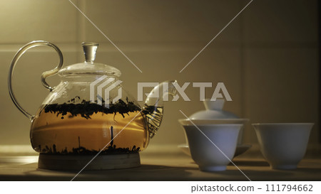 Teapot with fresh tea leaves in hot water and small cups on the kitchen table. Concept. Close up of glass transparent teapot with brewing black tea, concept of tea ceremony. Teapot with fresh tea leaves in hot water and small cups on the kitchen table. Concept. Close up of glass transparent teapot with brewing black tea, concept of tea ceremony. 111794662
