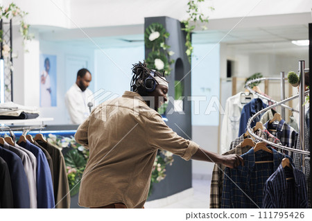 Cheerful african american man listening to music in headphones while browsing clothes in boutique. Happy smiling customer in earphones choosing casual shirt in shopping mall Cheerful african american man listening to music in headphones while browsing clothes in boutique. Happy smiling customer in earphones choosing casual shirt in shopping mall 111795426